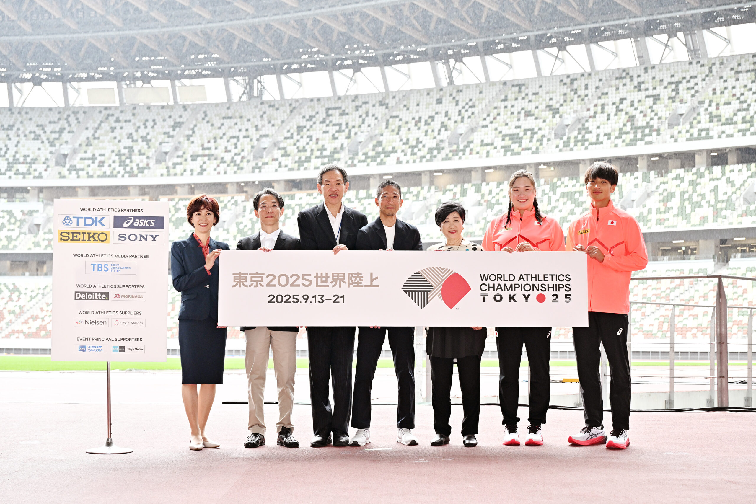 The group at the logo announcement event. From left, Yuko Arimori (The vice president of JAAF), Masahiko Kimura (The chairperson of Logo Design Selection Committee), 武市敬さん(東京2025世界陸上財団事務総長)、中川さん、小池百合子ささん(東京都知事)、北口榛花選手、橋岡優輝選手がロゴのパネルを持っている写真。