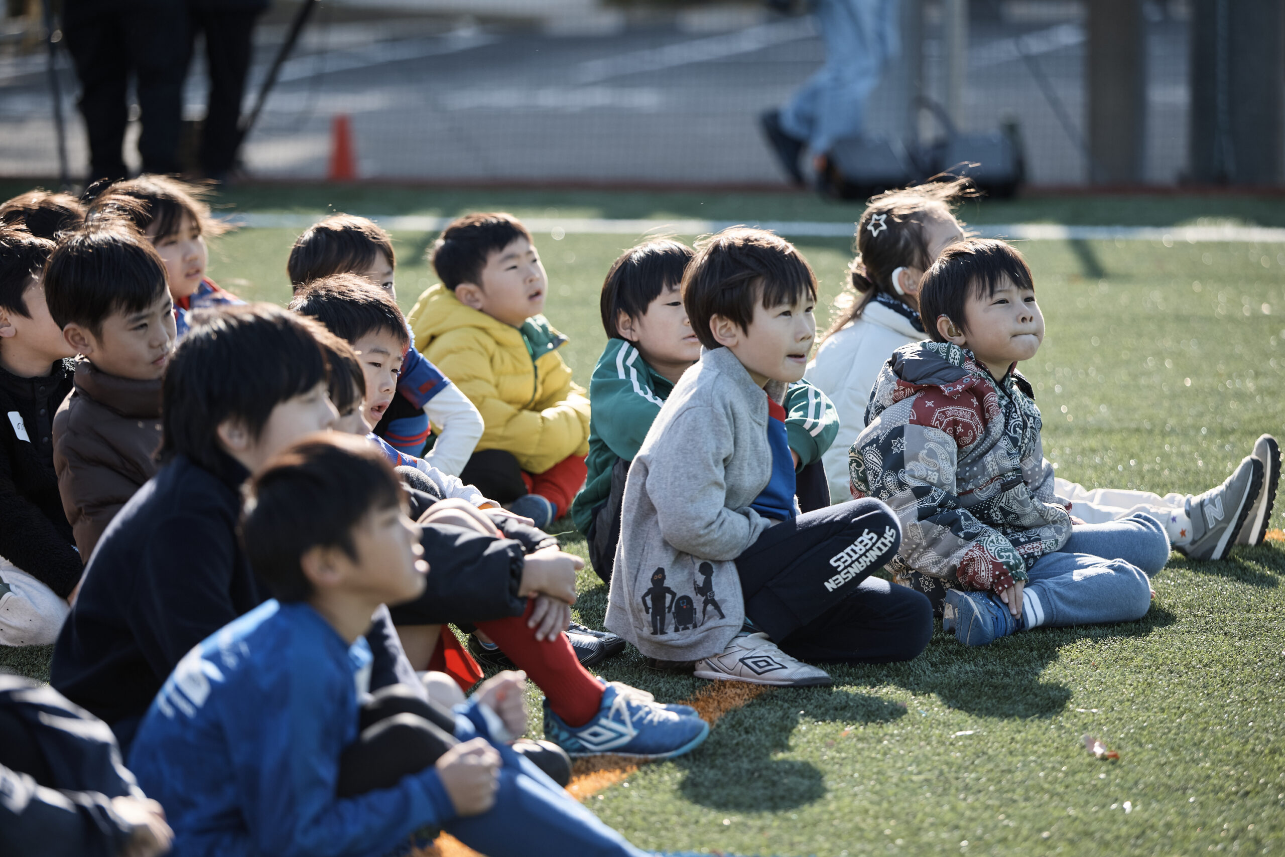 芝生の上で子供たちが選手の説明を真剣に聞いている写真