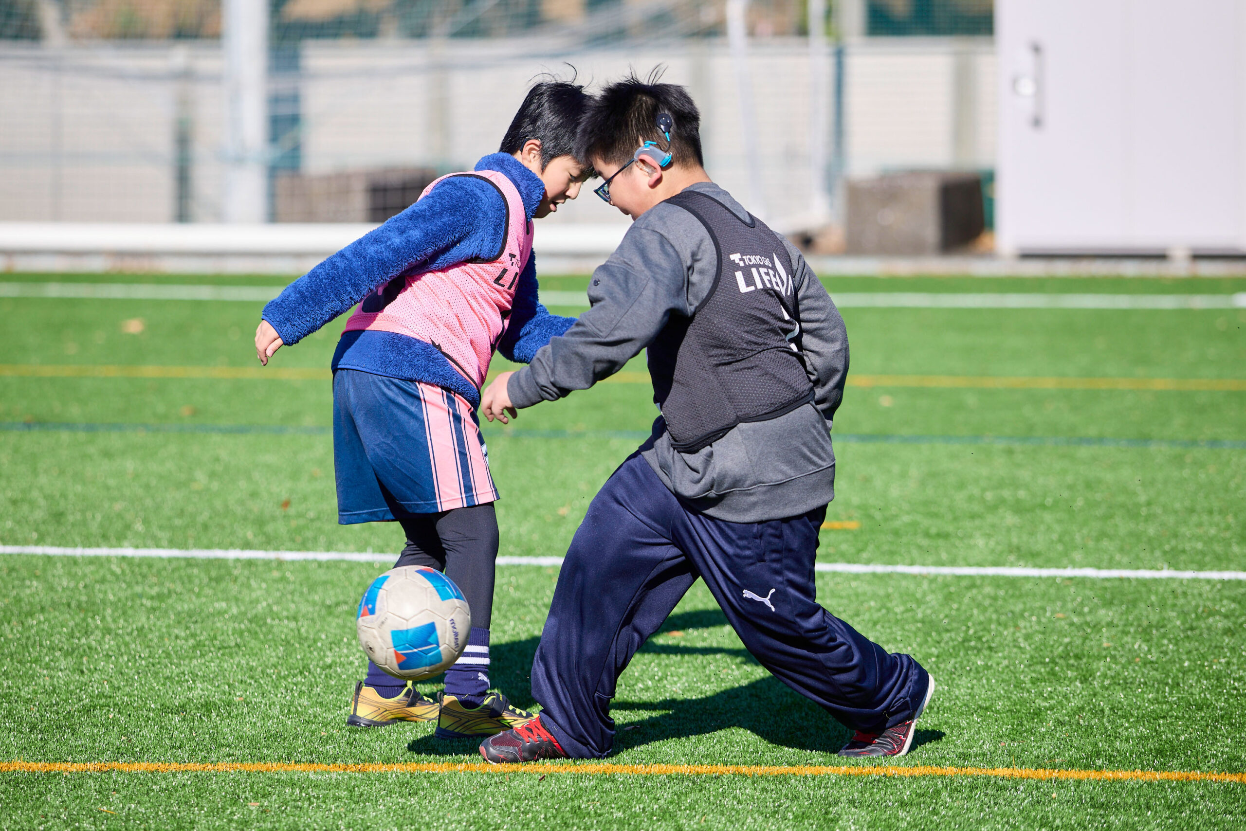 補聴器をつけた男の子とピンクのビブスをつけた男の子がボールを取り合っている写真